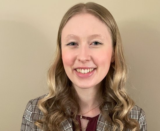 Abigail Smith, with long blond hair, wearing a flannel professional jacket, smiles and stands in front of a beige wall.