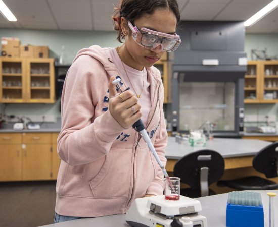 Yamilet Taveras ’27, a chemistry major wearing a pink hoodie and safety goggles, pours a substance into a beaker in the chemistry lab.