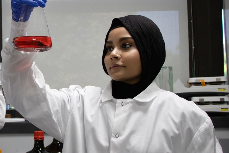 Nurhaliza Syukur, wearing a lab coat, holds up a beaker with red liquid in it.
