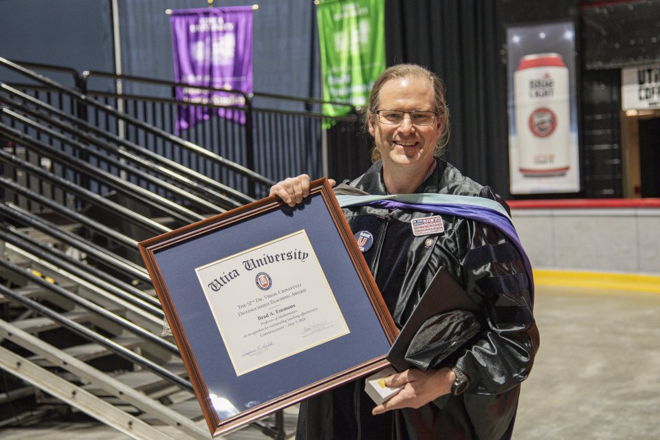Brad Emmons in commencement regalia, stands with the framed 2026 Crisafullli Award.